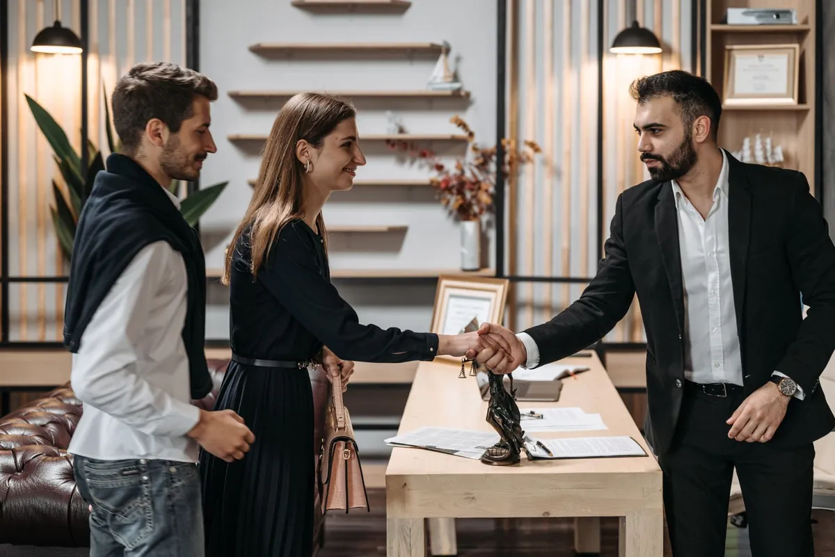 A couple shaking hands with a man in a suit across a desk with papers on it