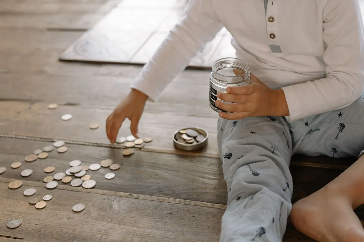 A child sitting on a wooden floor with a glass jar and coins scattered around