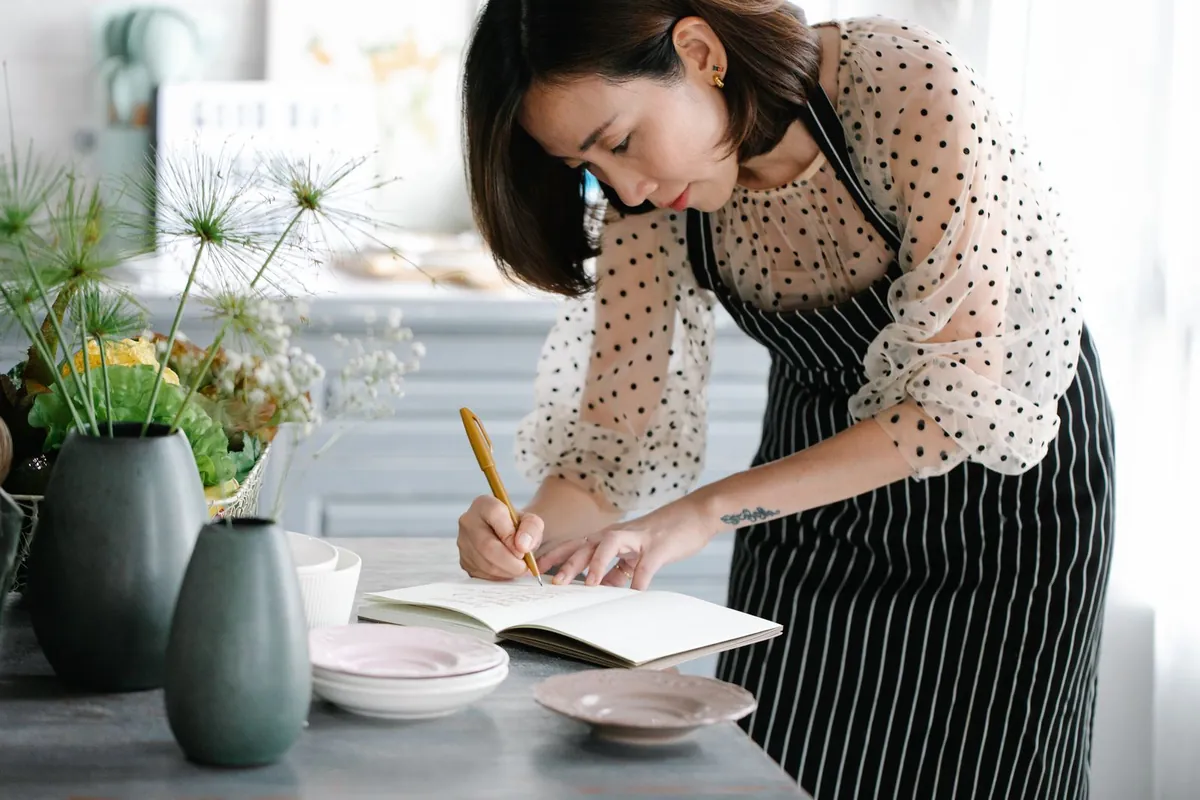 A woman in an apron writing in a notebook at a bright kitchen counter